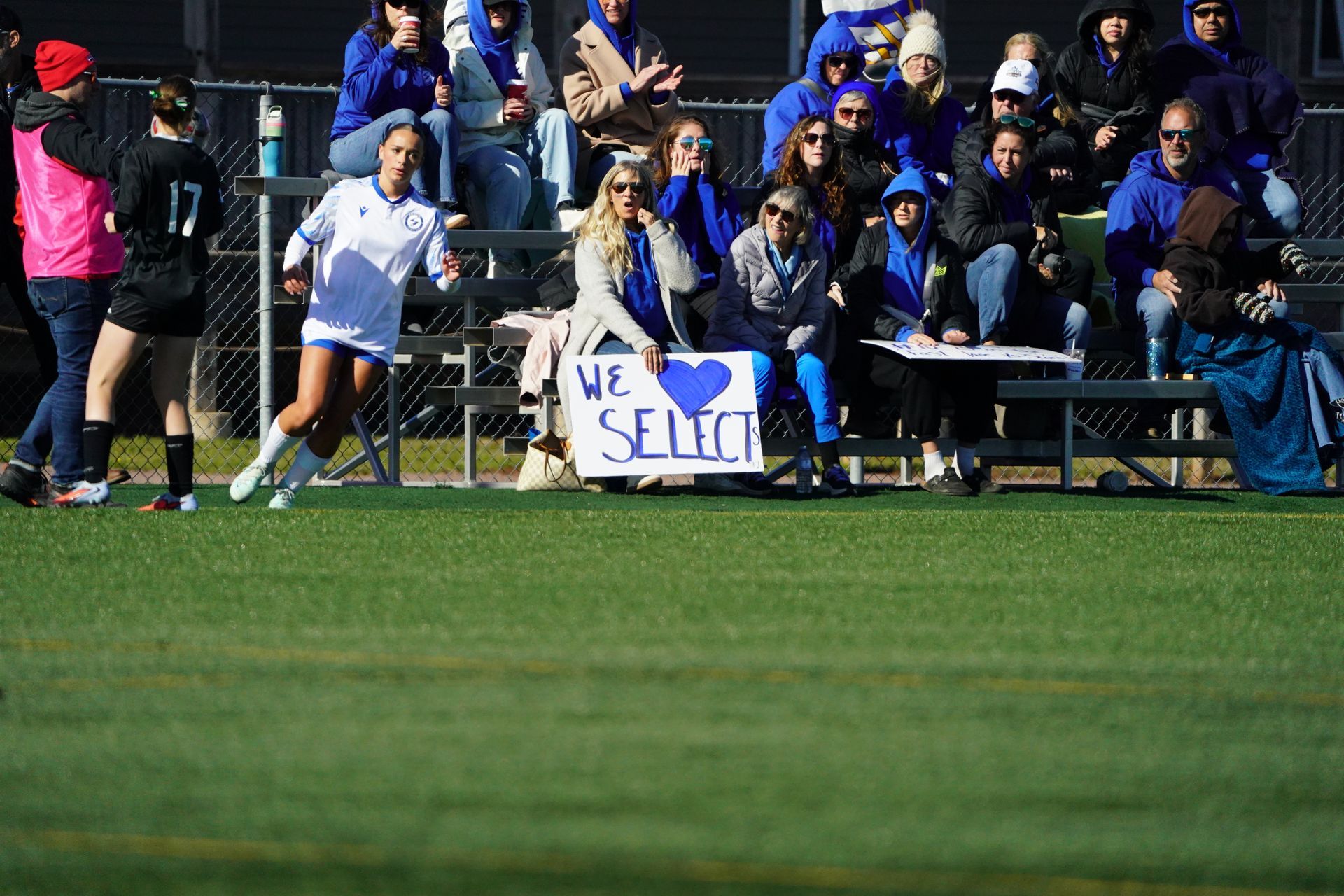 A soccer player in a white uniform runs past fans in bleachers holding a sign that reads 