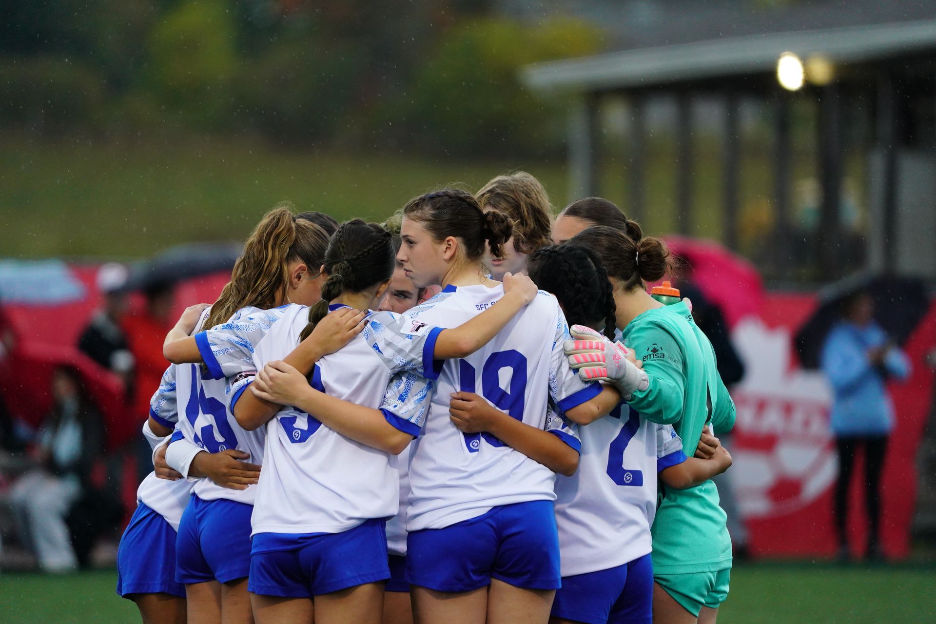 A soccer team in white and blue uniforms huddles together on a field with a goalkeeper in bright green.