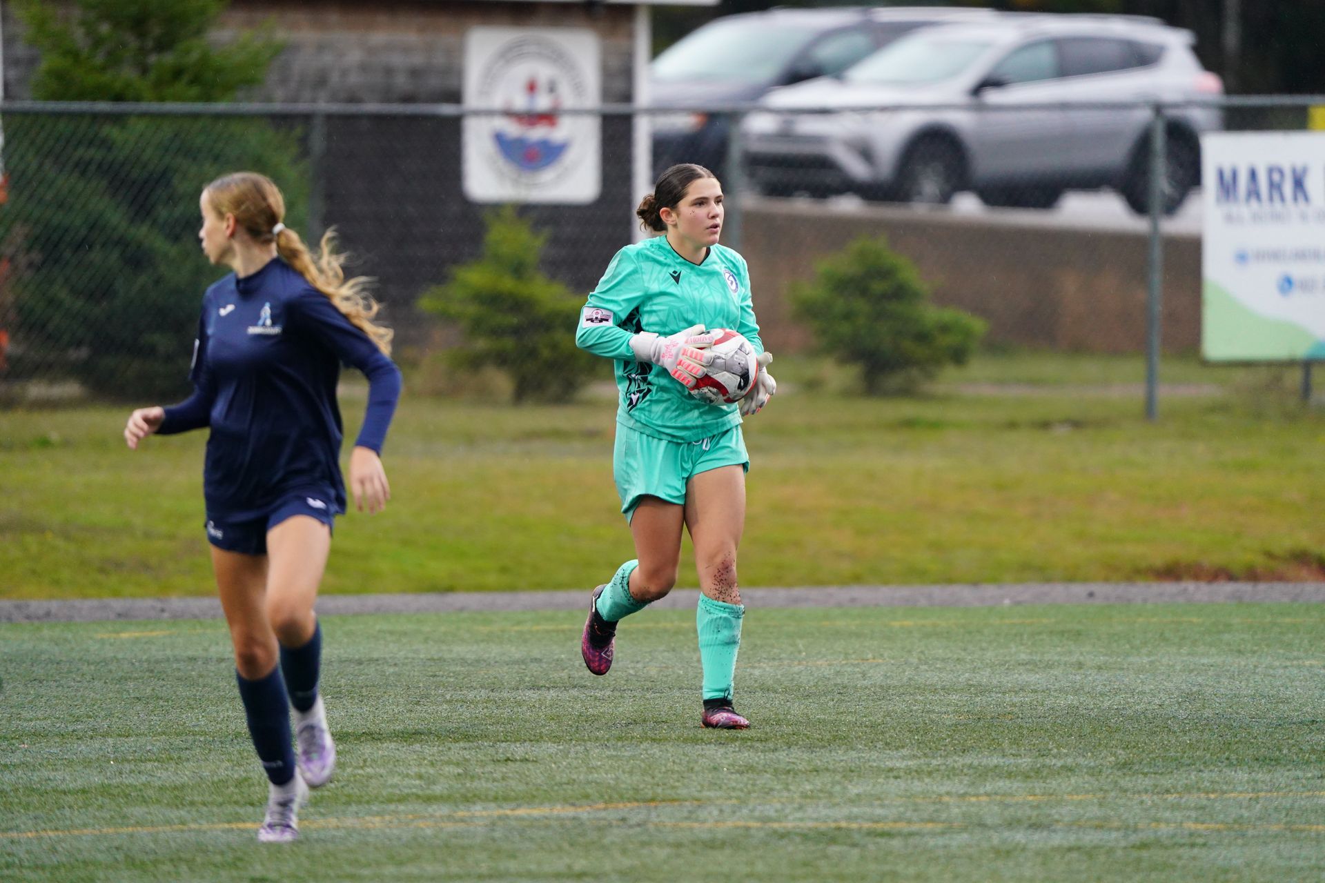 A soccer goalie in a bright green uniform holds a ball while running on a turf field near a teammate in dark blue.