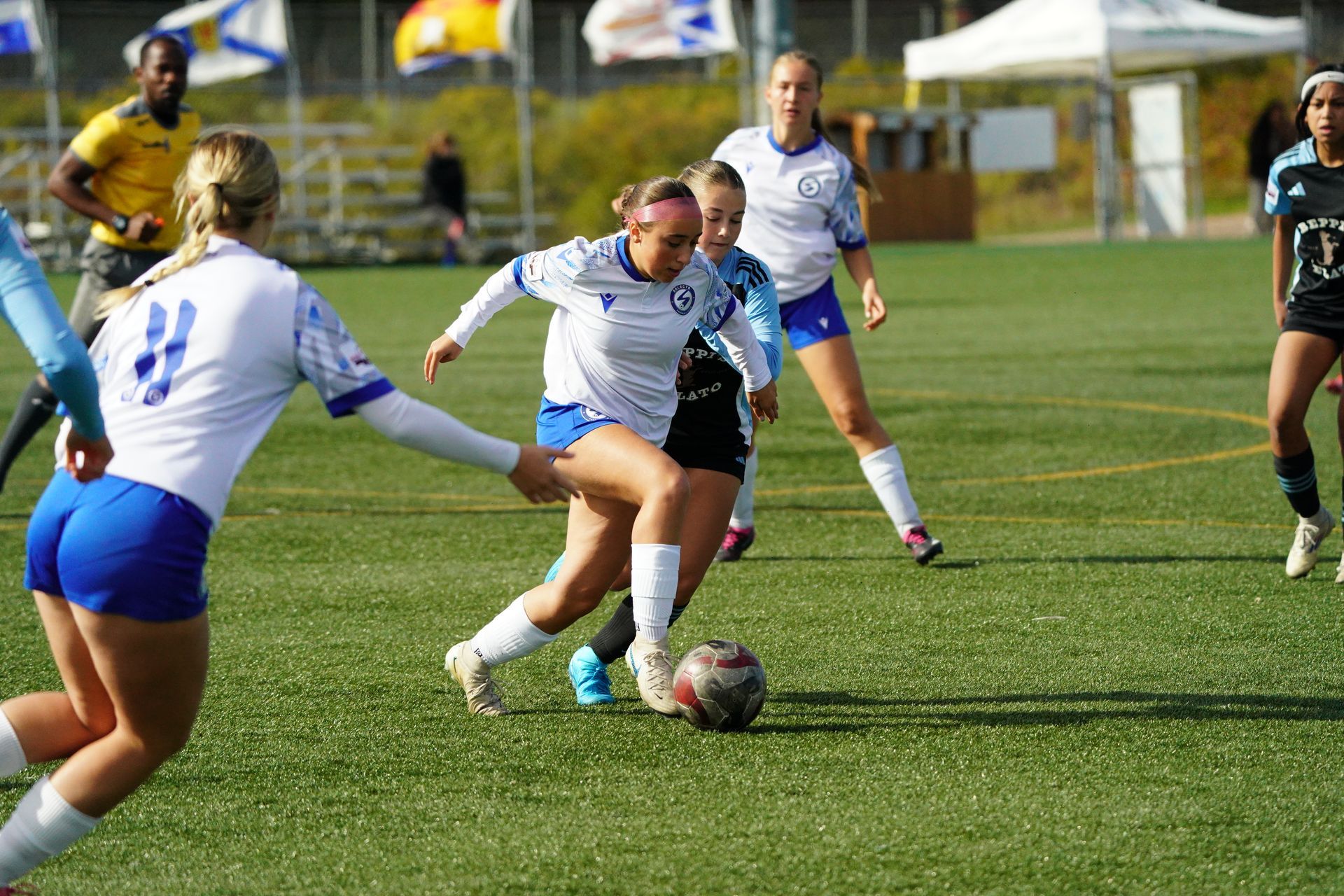 Soccer players in white and blue jerseys compete for the ball on a green field during an outdoor game.