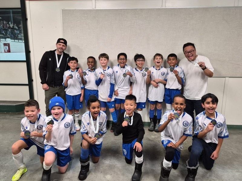 A soccer team in white and blue uniforms standing with medals and two coaches indoors against a plain wall.