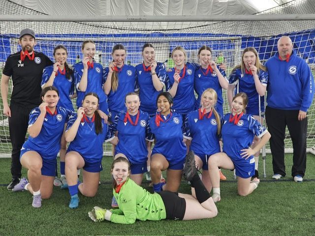 A soccer team in blue uniforms and medals posing for a photo on an indoor field with two coaches.