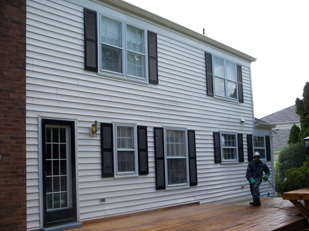 A man is standing on a deck in front of a white house with black shutters