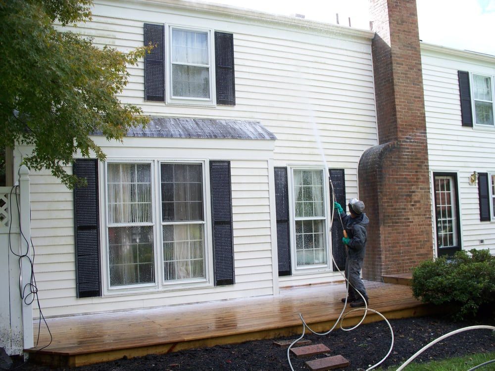 A man is washing the side of a white house with black shutters.