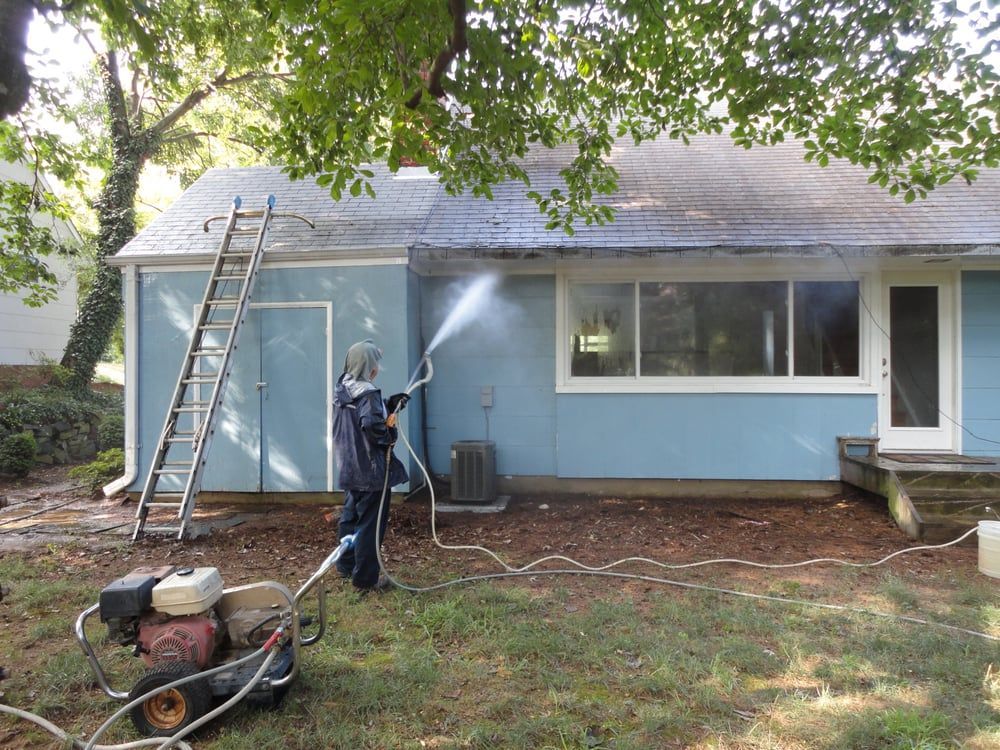 A man is cleaning the roof of a blue house with a pressure washer.