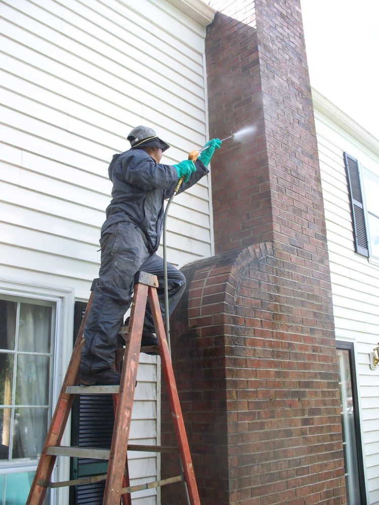 A man is standing on a ladder cleaning a brick chimney.