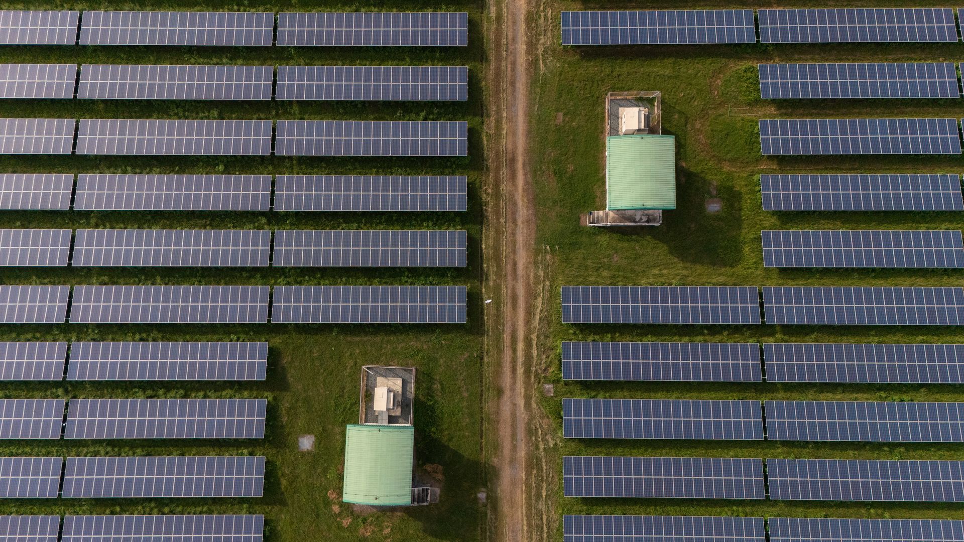Aerial view of a solar panel farm with rows of panels, dirt pathway, and green-roofed structures.