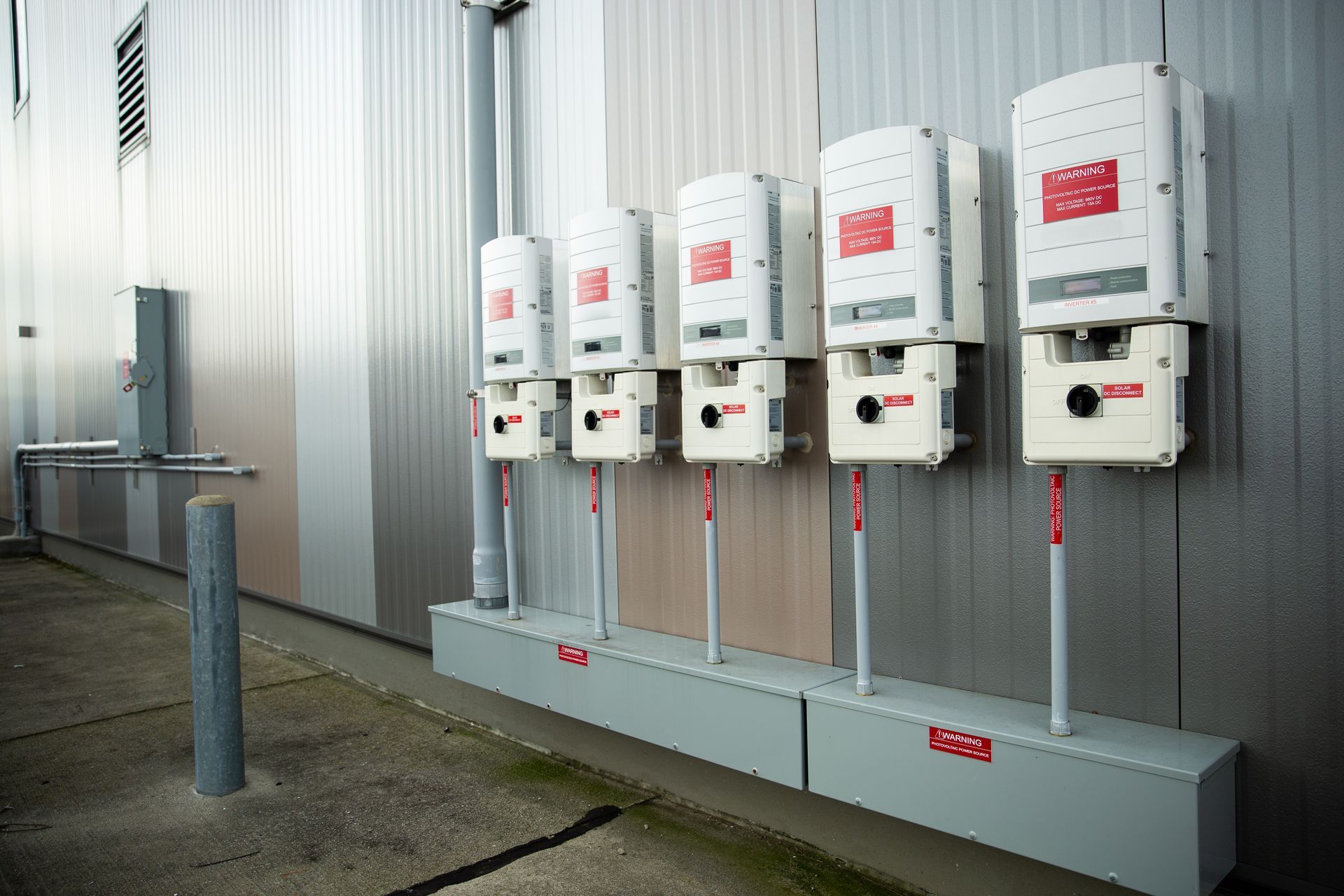 White solar inverters mounted on a wall with red warning labels and gray conduit boxes.