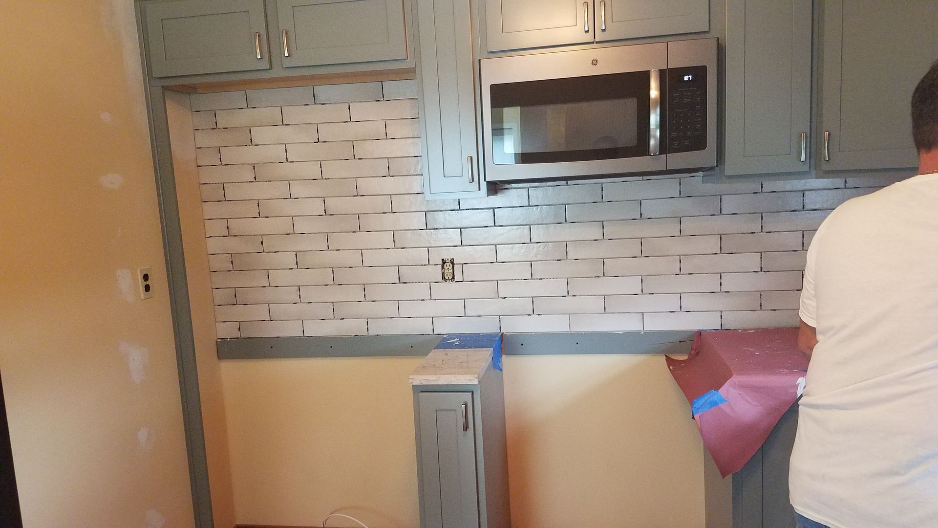 A man is standing in a kitchen with a microwave and a brick wall.