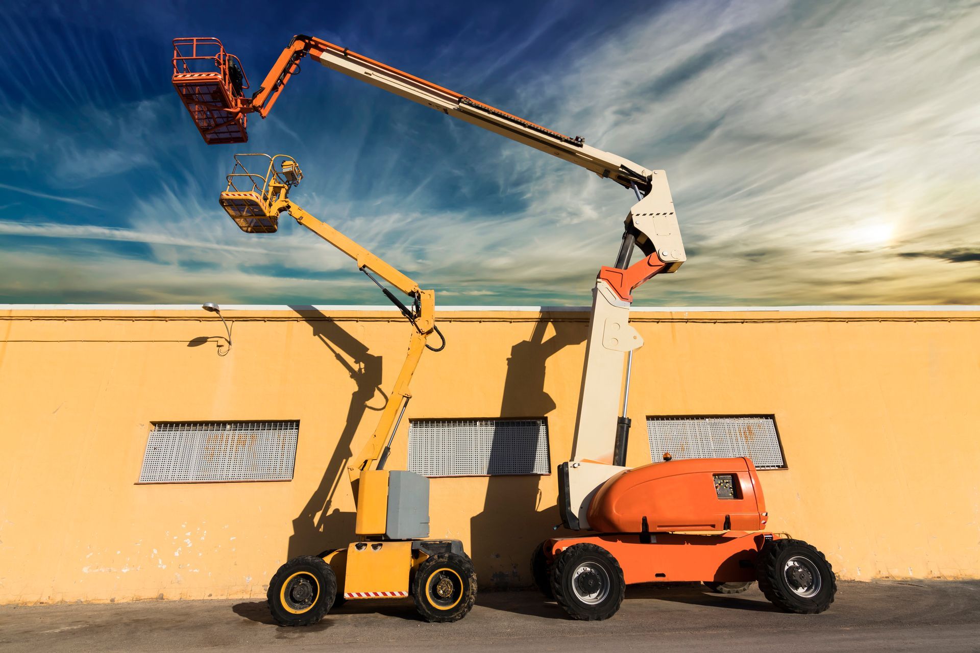 Two aerial lifts, one yellow, one orange, against a yellow building and blue sky.