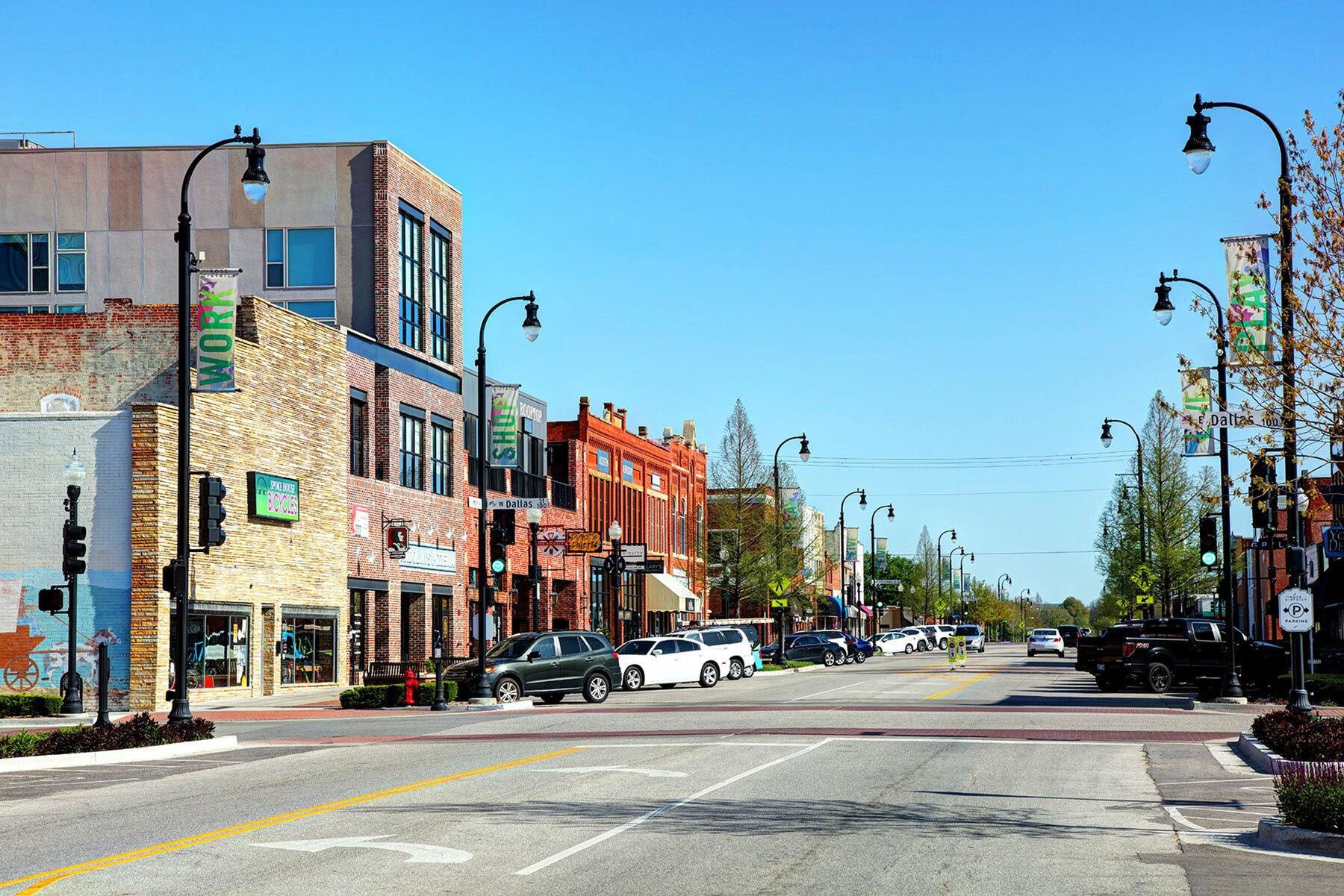 A sunlit downtown street with historic brick buildings, streetlights, parked cars, and clear blue skies.
