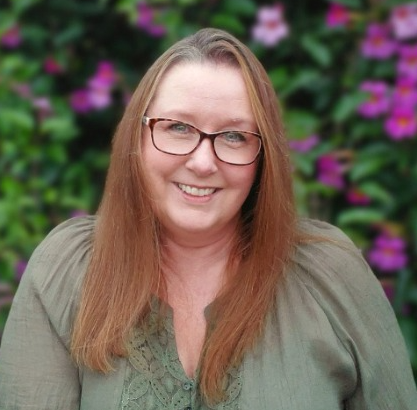 Smiling woman with glasses in a green blouse, standing before a blurred flower garden.