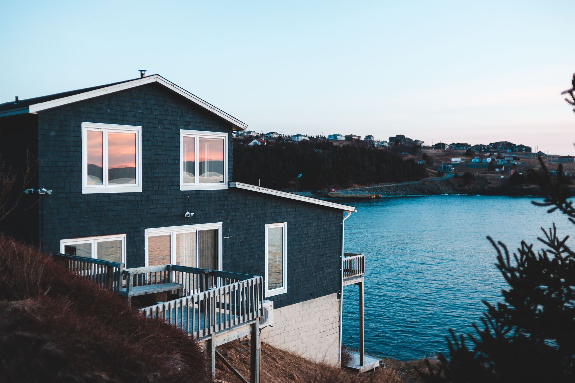 Dark gray house with lit windows beside calm blue water at dusk
