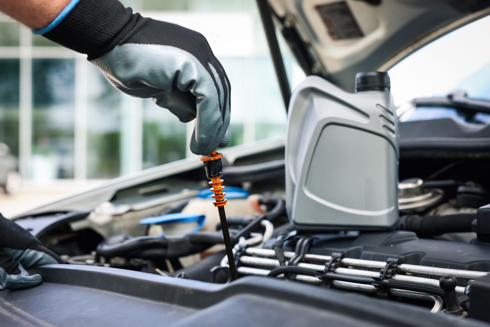 Gloved hands checking car oil dipstick, near open engine and oil container.