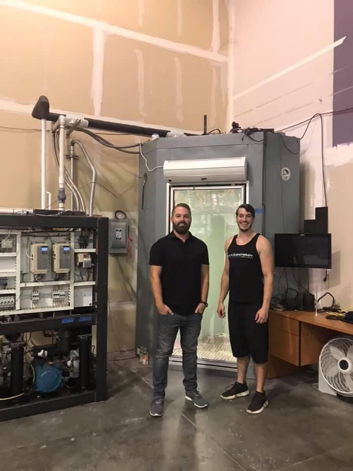 two men are standing in front of a large cryotherapy machine in a room .