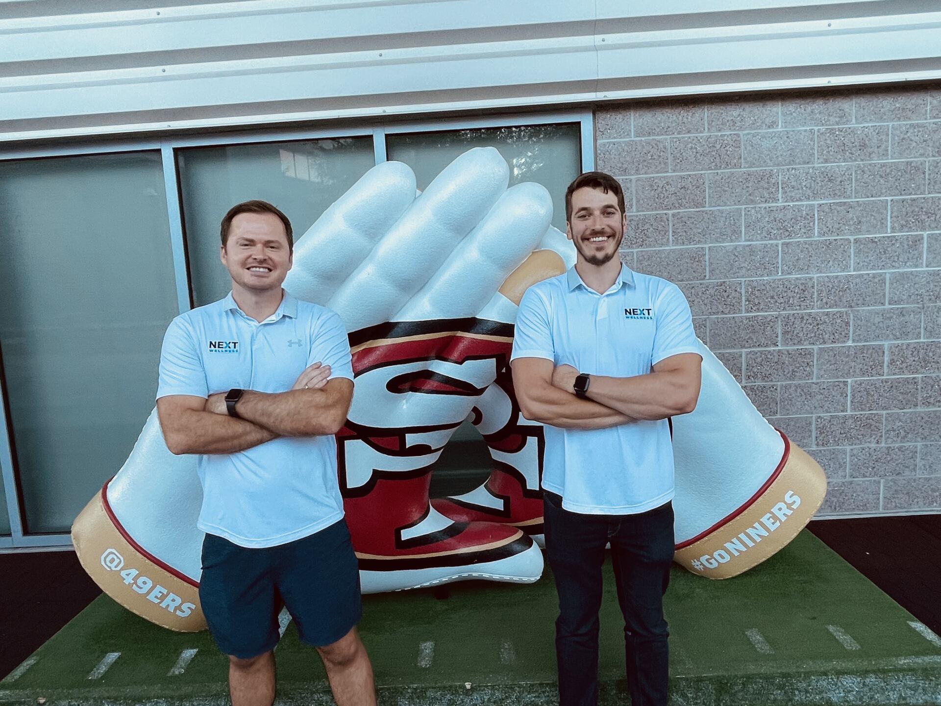 two men who just sold equipment are standing next to each other in front San Francisco 49ers logo .