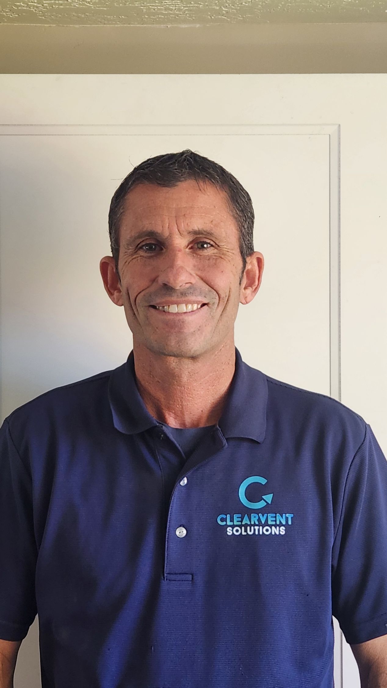 Man standing indoors in a navy polo shirt, smiling against a light wall.