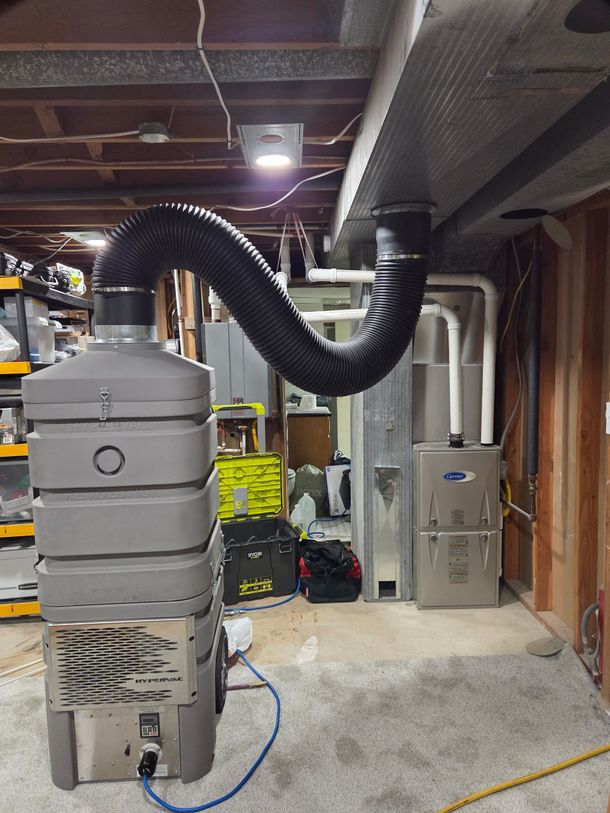 Basement utility room with a large gray HVAC/furnace unit, black ducting, and exposed ceiling pipes.