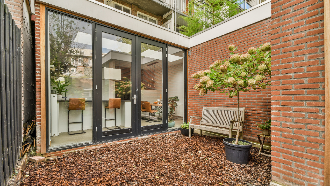 Small courtyard with glass doors, brick walls, a bench, and a tree on wood chips.