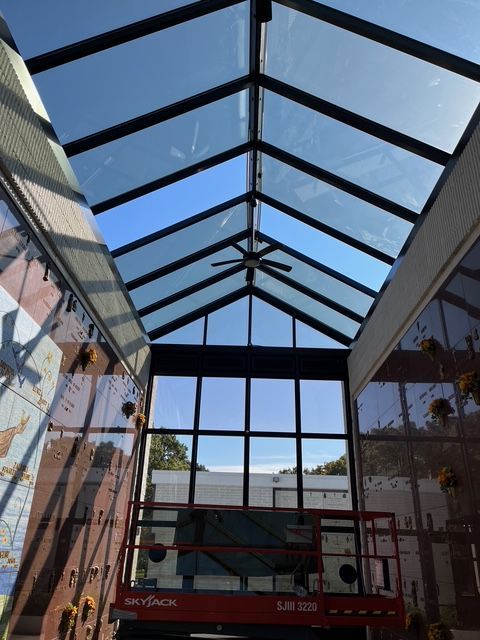 Interior with angled glass ceiling, black frame, and a scissor lift visible at the base of the structure.