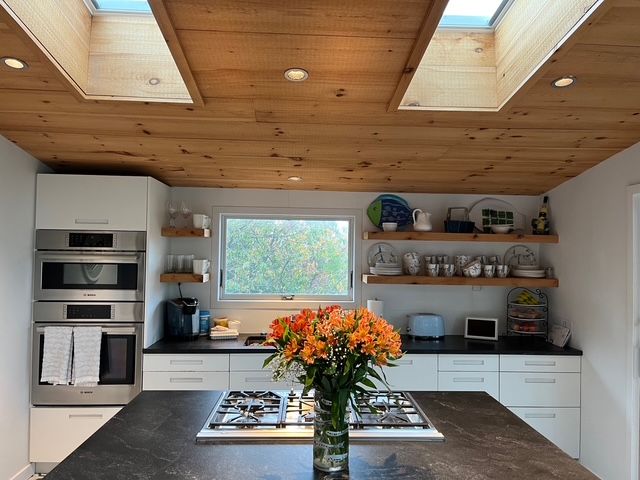 Kitchen with wood ceiling, skylights, white cabinets, island with stovetop, and orange flowers.