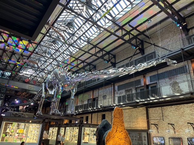 A large, reflective dinosaur sculpture hangs inside a multi-story building with a glass ceiling.
