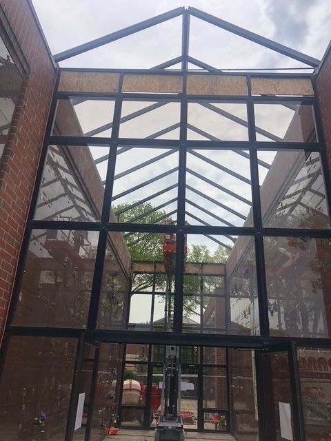 Large glass and steel window installation between brick structures; a person works on a lift.