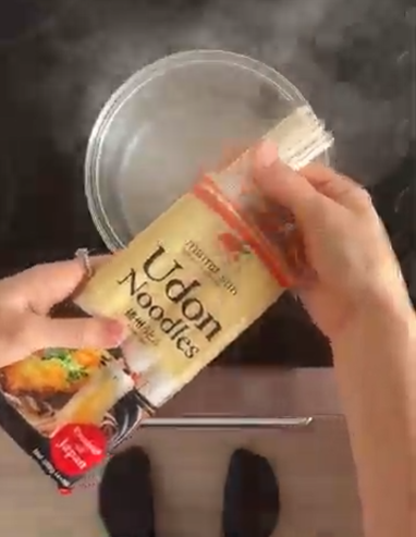 Hands pouring dried Udon noodles from a package into a pot of boiling water on a stove.