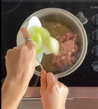 A person’s hands pouring sliced onions from a white plate into a metal pot containing browned meat pieces on a stovetop.