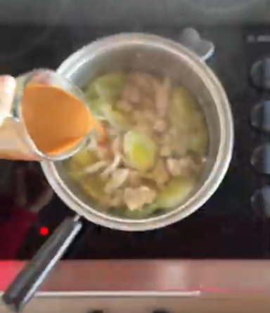 A hand pours a light brown liquid into a metal pot on a black stovetop containing chopped leeks and pieces of meat.