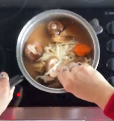 Hands stir sliced onions and whole mushrooms in a pot of brown liquid on a stovetop.