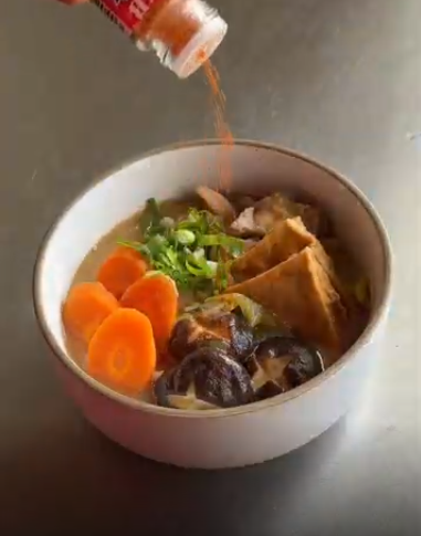 A hand sprinkles red seasoning over a bowl of soup with sliced carrots, mushrooms, tofu, and green onions.