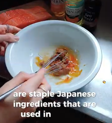 Hands whisk miso paste and egg in a white bowl, with bottles of mirin, sake, and soy sauce and salmon nearby.