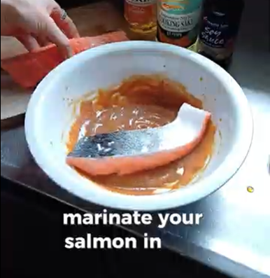 A person places a salmon fillet into a bowl containing a smooth, light-brown marinade.