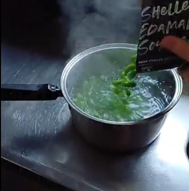 Hand pouring shelled green edamame soybeans into a pot of boiling water on a stainless steel surface.