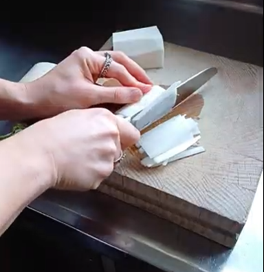 A person uses a kitchen knife to thinly slice a white vegetable on a wooden cutting board.