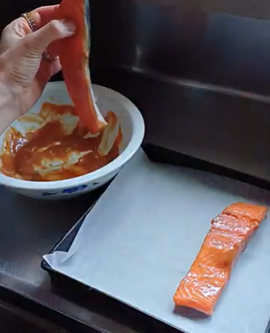 A hand dips a piece of raw salmon into a bowl of sauce next to a baking sheet with another piece of salmon.