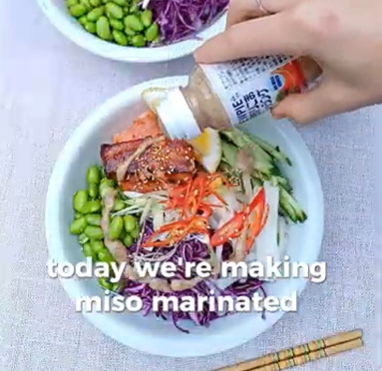 A hand squeezes dressing from a bottle onto a vibrant poke bowl with edamame, red cabbage, cucumber, and miso salmon.