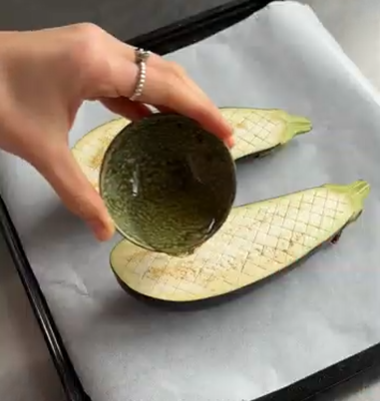 Hand holding a halved eggplant over two scored avocado halves on a baking tray.