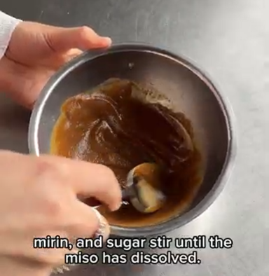 Hand mixing miso, mirin, and sugar in a small metal bowl on a countertop.