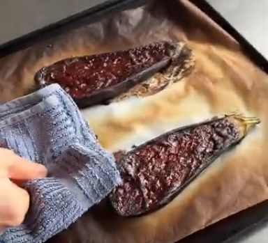 Hand wiping a baking tray with two dark roasted eggplants on parchment paper