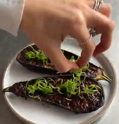 Hand placing green garnish on two grilled eggplants on a white plate