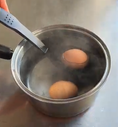 Two eggs simmering in a metal pot with a ladle on the left, steam rising from the water.