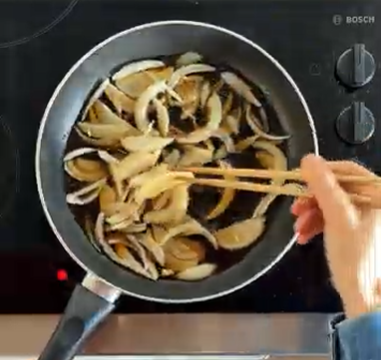 Hand stirring sliced onions in a skillet on a stovetop with chopsticks