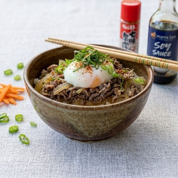 Beef rice bowl topped with a soft egg and green onions, with chopsticks and sauce bottles nearby