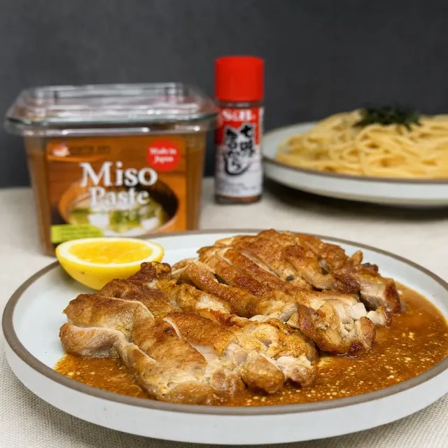 Plate of chicken in brown miso sauce with lemon wedge, miso paste jar, seasoning, and noodles in background