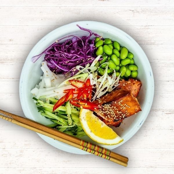 A poke bowl with glazed salmon, edamame, shredded red cabbage, julienned cucumber, chili slices, and a lemon wedge.