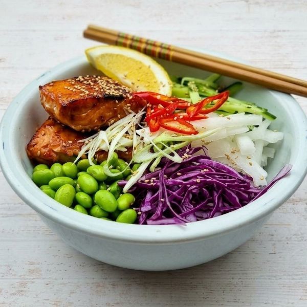 A poke bowl with glazed salmon, edamame, shredded red cabbage, julienned cucumber, chili slices, and a lemon wedge.