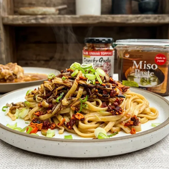 Plate of udon noodles topped with crunchy garnish, with miso paste and crunchy garlic jars in the background