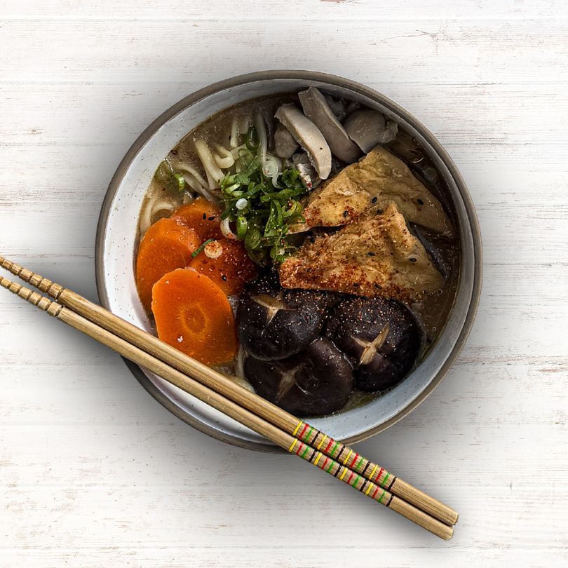 Bowl of noodle soup with vegetables and chopsticks on a white table.